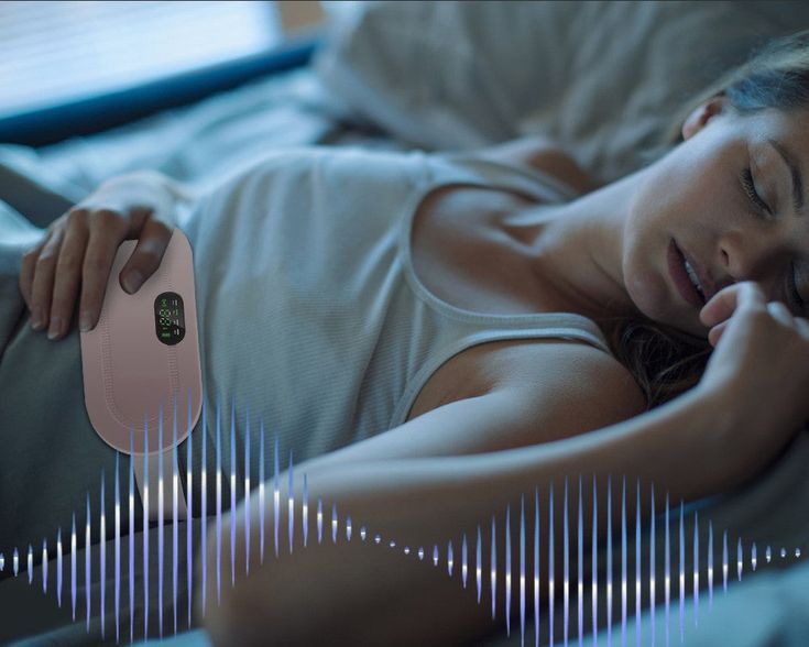 Woman lying on a bed with a pink electronic device on her stomach, possibly an abdominal massager.