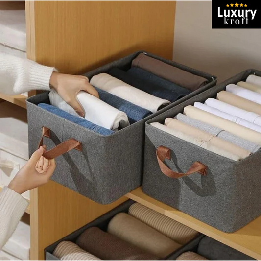 Gray storage bins with handles containing folded clothes inside a wooden wardrobe, branded 'Luxury Kraft'.